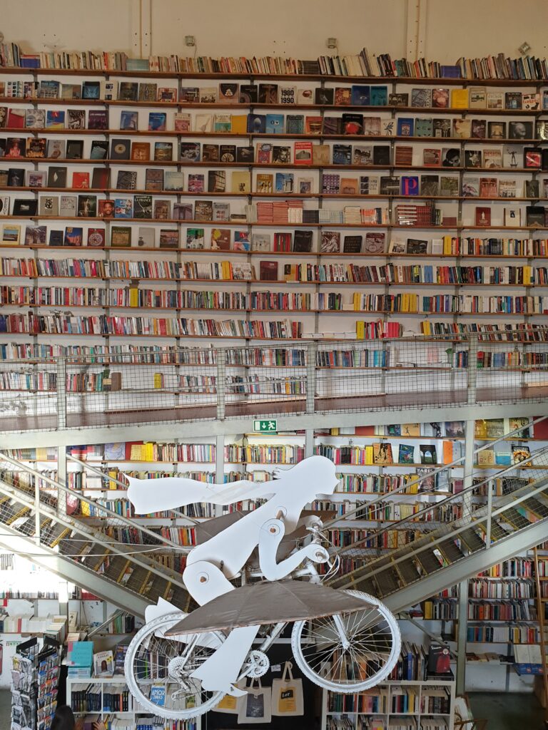 Una foto di una parete della libreria Ler Devagar colma di Libri. In primo piano di vede una decorazione sospesa in aria che rappresenta una bambina su una bicicletta