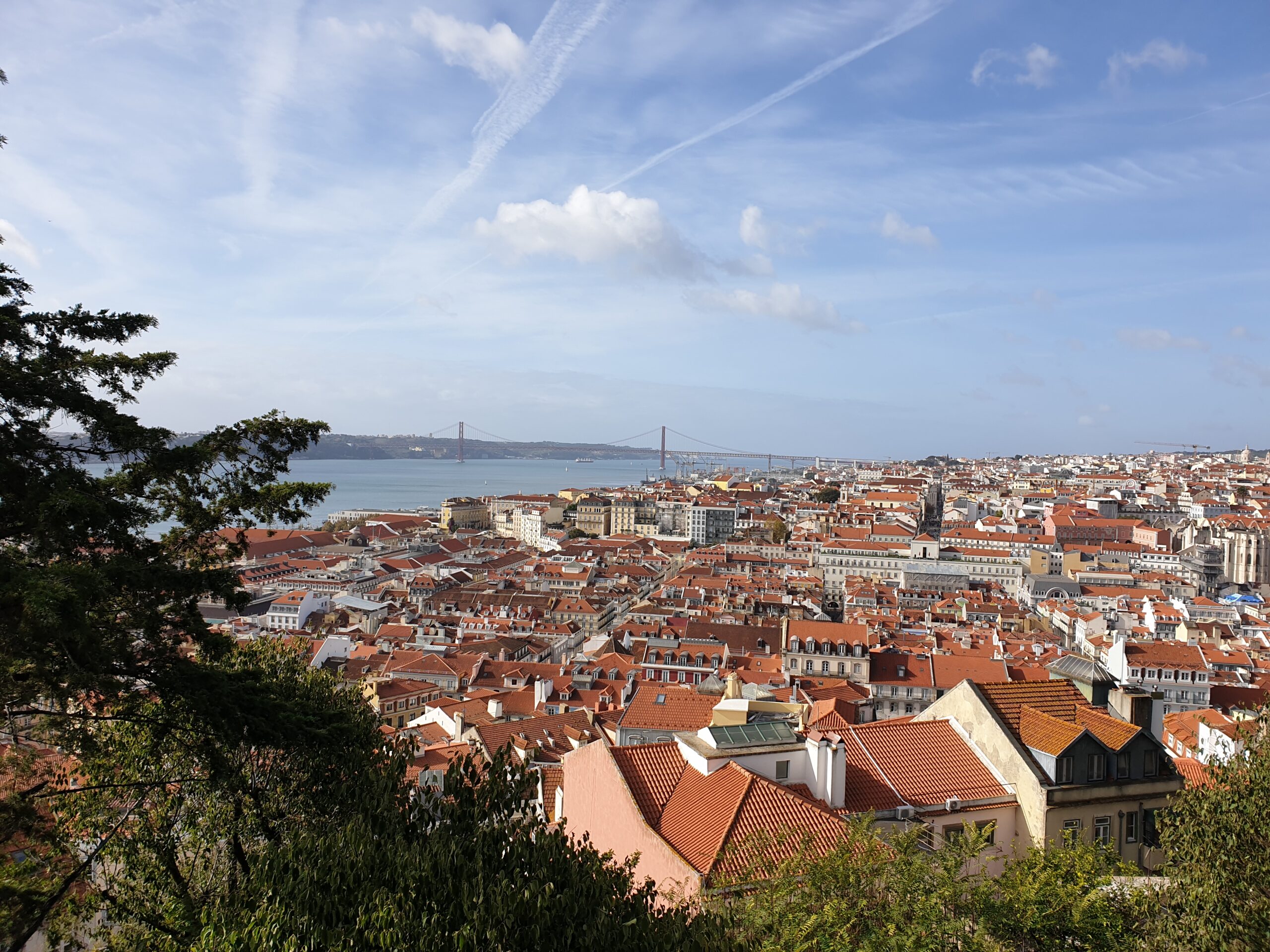 In foto si vede Lisbona dall'alto: spiccano i tetti rossi, il fiume e il ponte rosso ad archi che ricorda quello di San Francisco