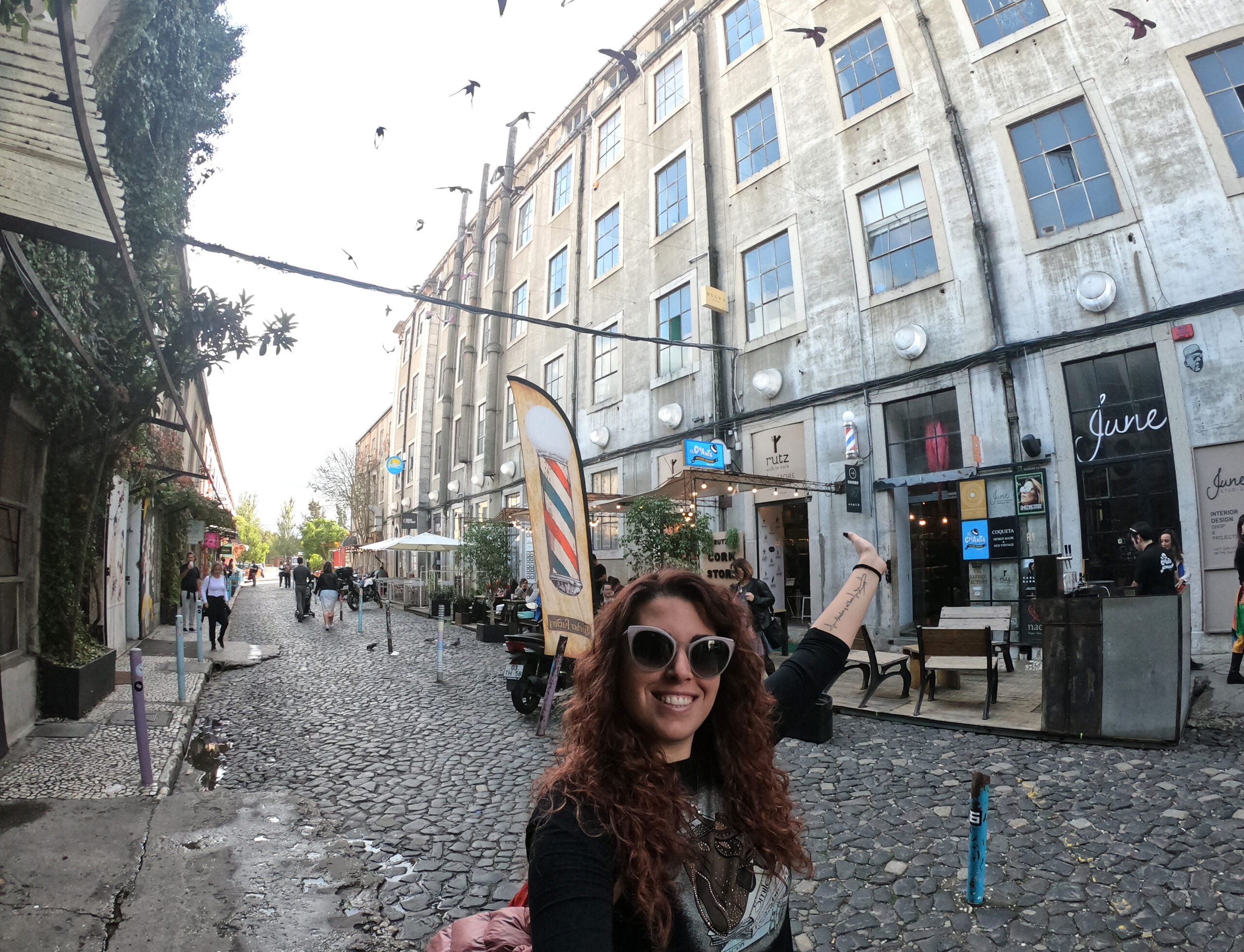 Carlotta sorridente e con un braccio alzato verso l'alto nel quartiere LX factory di Lisbona davanti ad un ex edificio industriale riconvertito in un pub