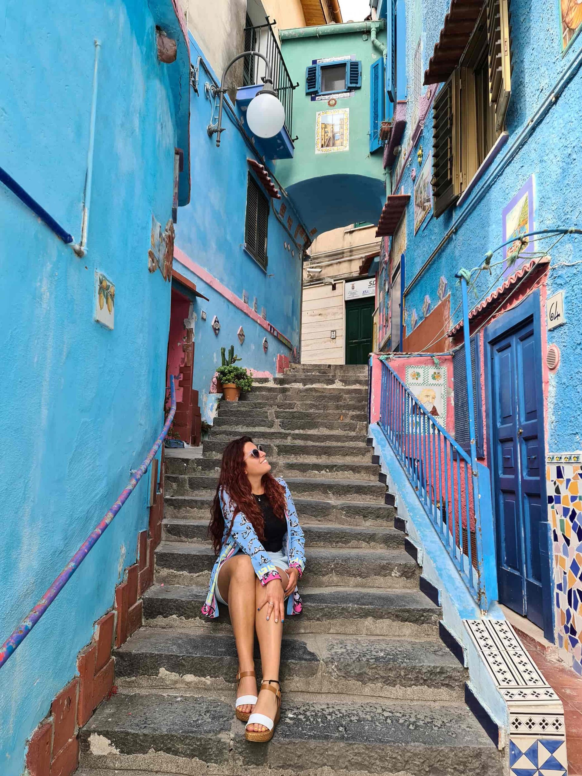 Carlotta seduta sulle scale, circondata da due file di edifici tutti azzurri e con mattonelle di ceramica colorata appese alle pareti.
