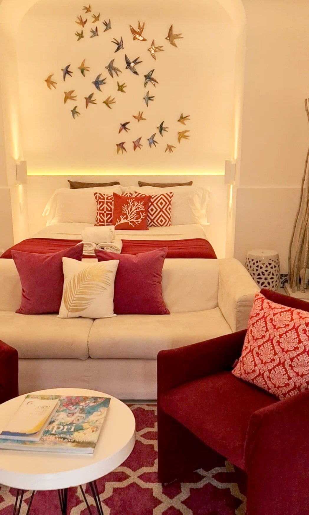 Una foto di una suite a Capri, con un grande letto matrimoniale con tanti cuscini rossi, appesi alla parete alle spalle del letto tante rondini in ceramica, mentre in primo piano c'è una poltroncina di velluto e un tavolinetto bianco con alcune riviste sull'isola