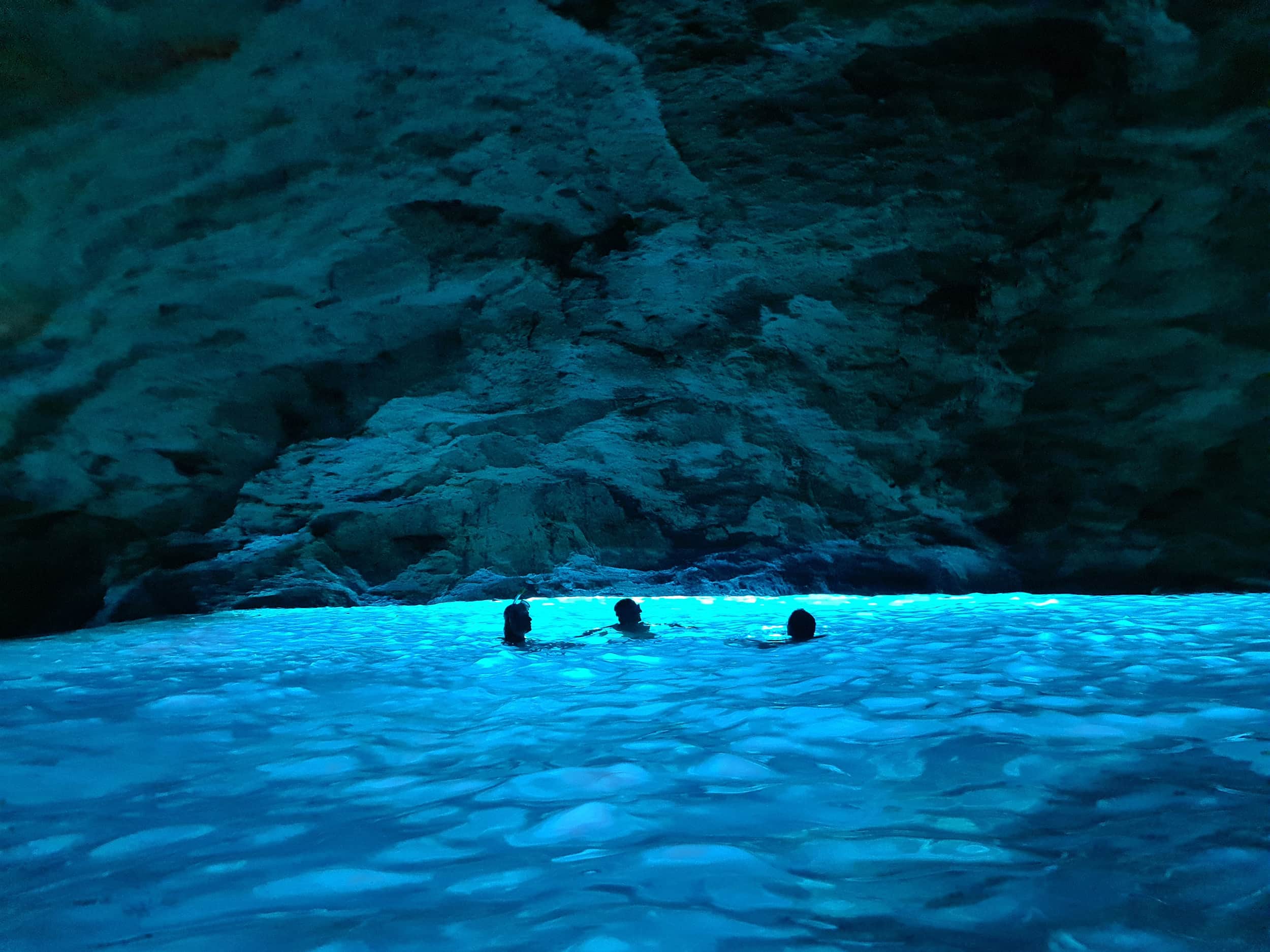 In foto la grotta Verde, un'ampia cavità illuminata di azzurro-verde dalla luce solare che filtra dal basso. Nell'acqua ci sono tre persone in controluce, che sembrano fare il bagno nella luce azzurra; è una delle grotte del Salento più belle.