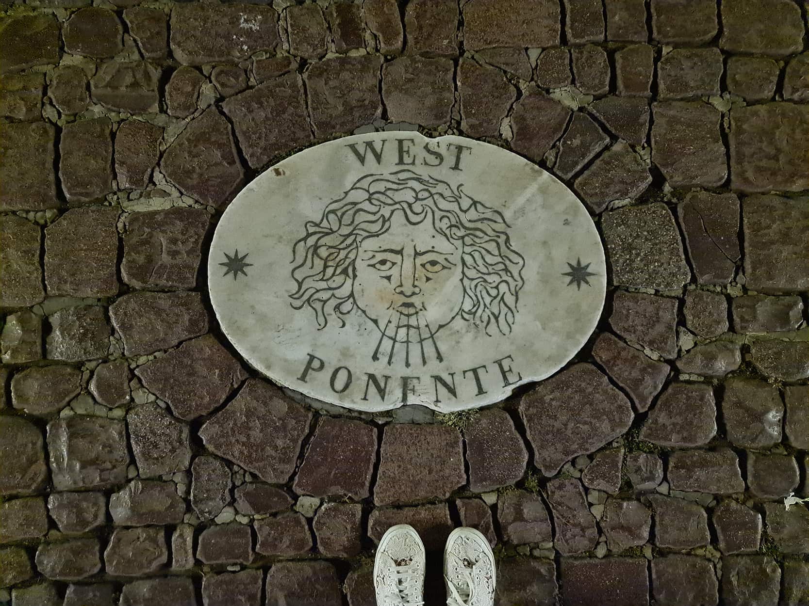Uno dei bassorilievi presenti in piazza San Pietro, ovvero una piastrella sul pavimento di san pietrini, raffigurante un angelo che soffia. Sul bassorilievo c'è scritto west ponente.