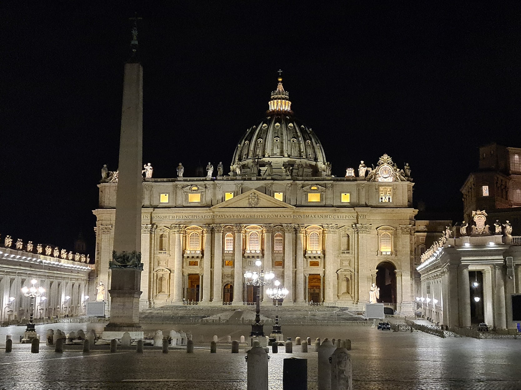 Una foto della Basilica di San Pietro di sera, illuminata e vuota, con l'obelisco in primo piano e il cupolone in bella vista.