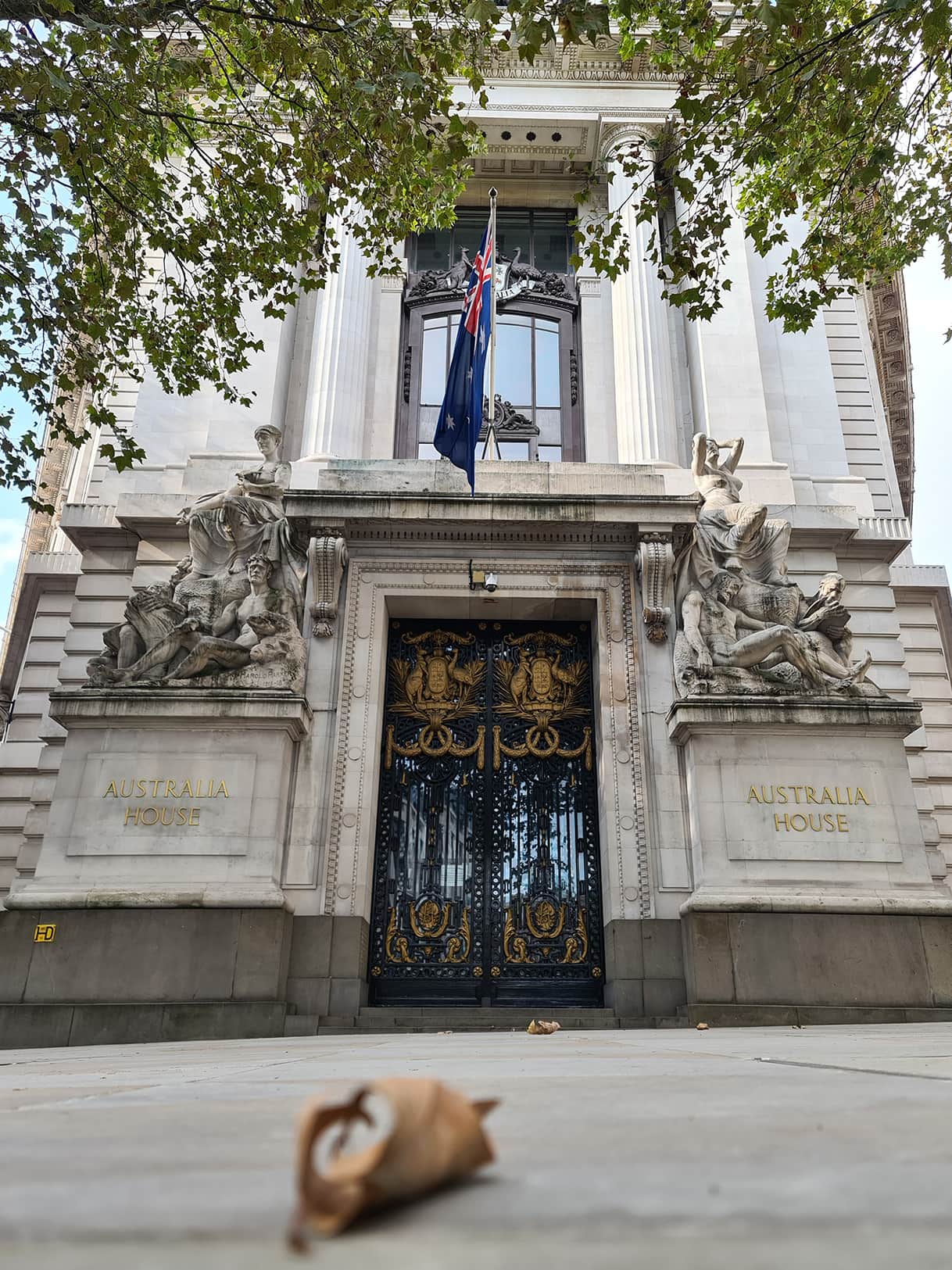 Si vede una foglia ingiallita per terra sfocata, e la facciata della Australia House, tutta bianca con la scritta dorata sul muro e una grande porta nera.