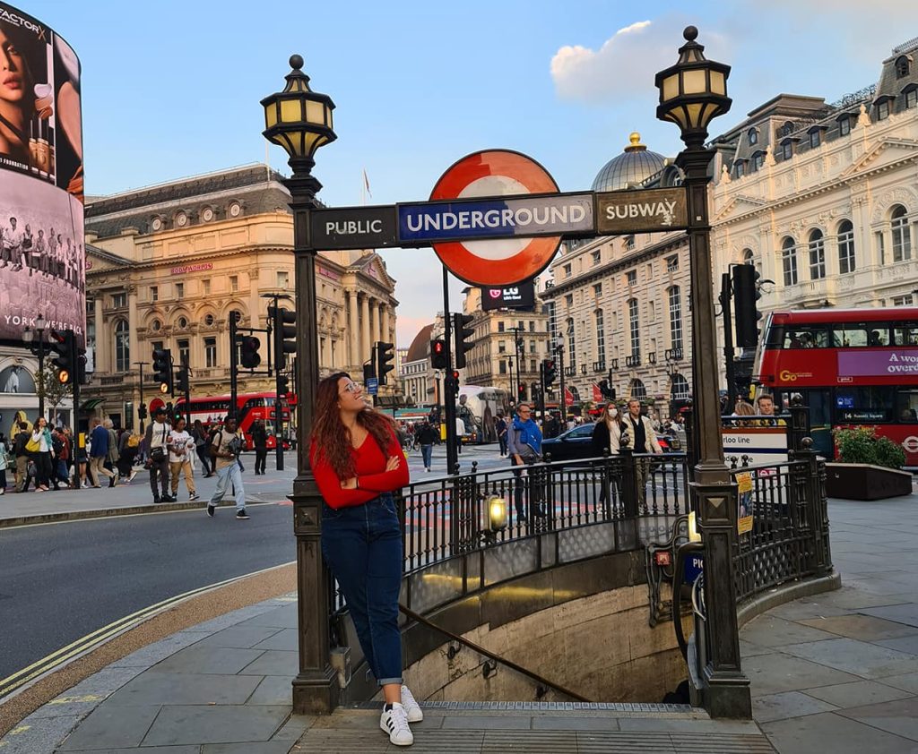 Carlotta, con indosso un paio di jeans ed una maglietta rossa, appoggiata ad un lampione situato all'ingresso delle scalette che portano ad una fermata metro a Piccadilly Circus