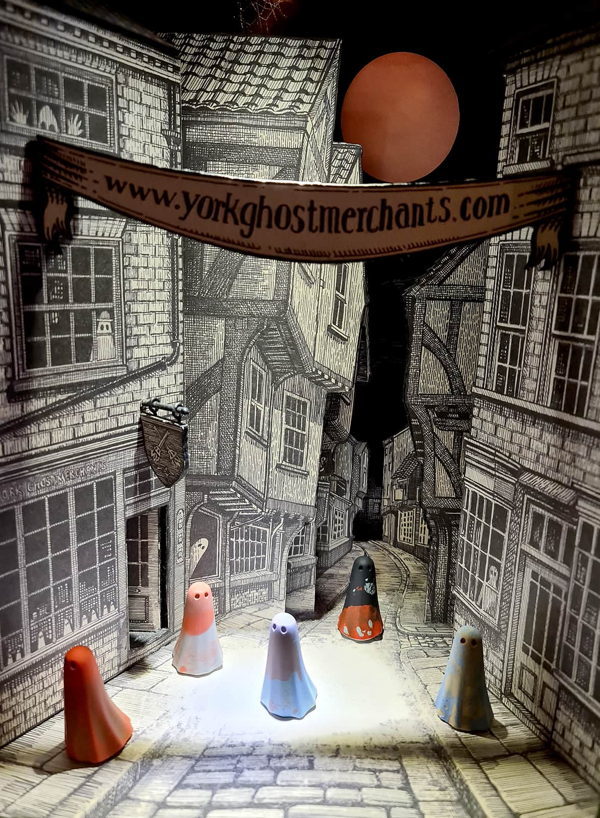 In foto si vedono 5 statuine di piccoli fantasmi con il lenzuolo colorato addosso, sullo sfondo di un cartonato che riprende il centro storico di York