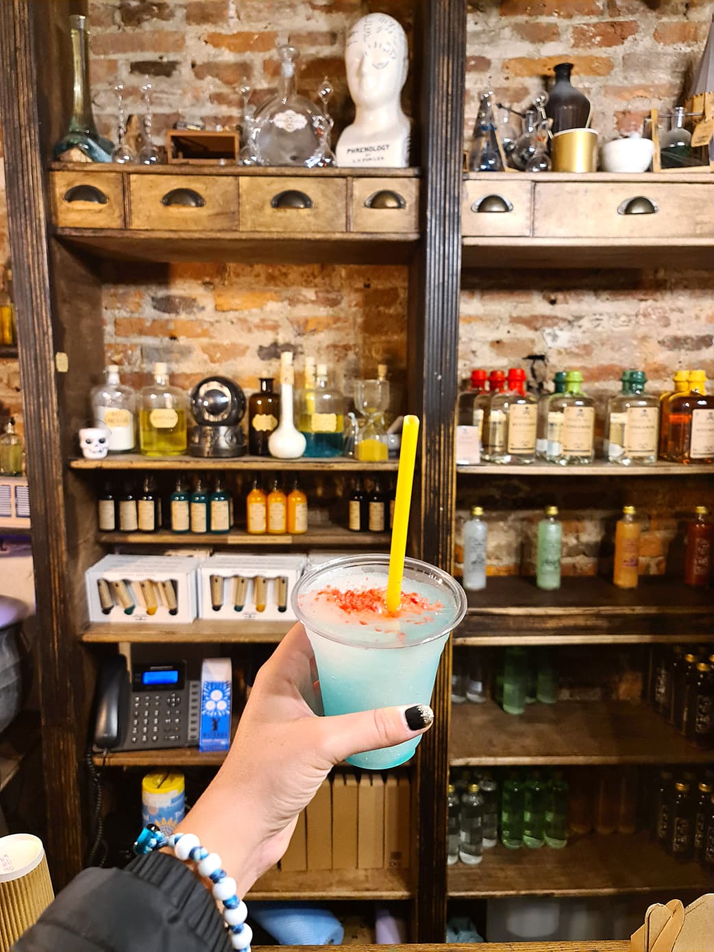 In foto si vede la mano di Carlotta mentre regge un bicchiere contenente una specie di cocktail colorato, sullo sfondo di una parete ricoperta di boccette e pozioni magiche