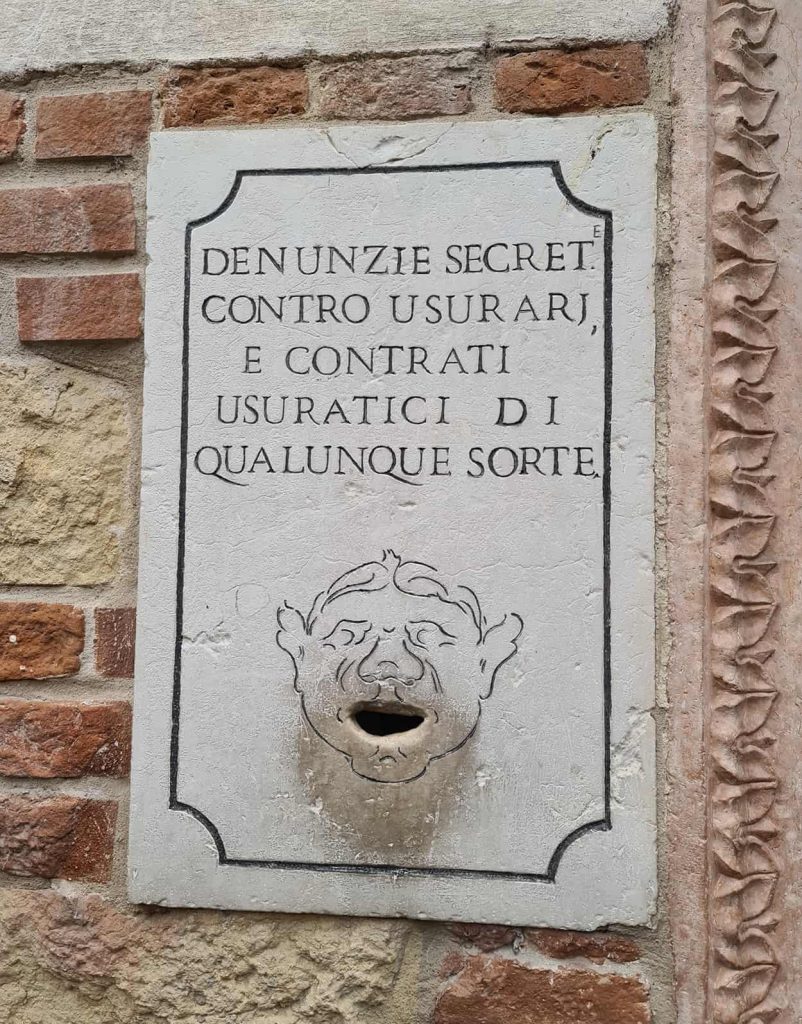 In foto si vede una targa in pietra bianca con una scritta e una apertura a forma di bocca al centro