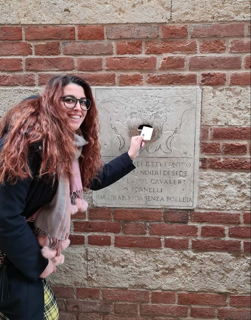 Carlotta sorridente, mentre finge di inserire un foglietto di carta all'interno della bocca sul muro del Palazzo della Ragione a Verona