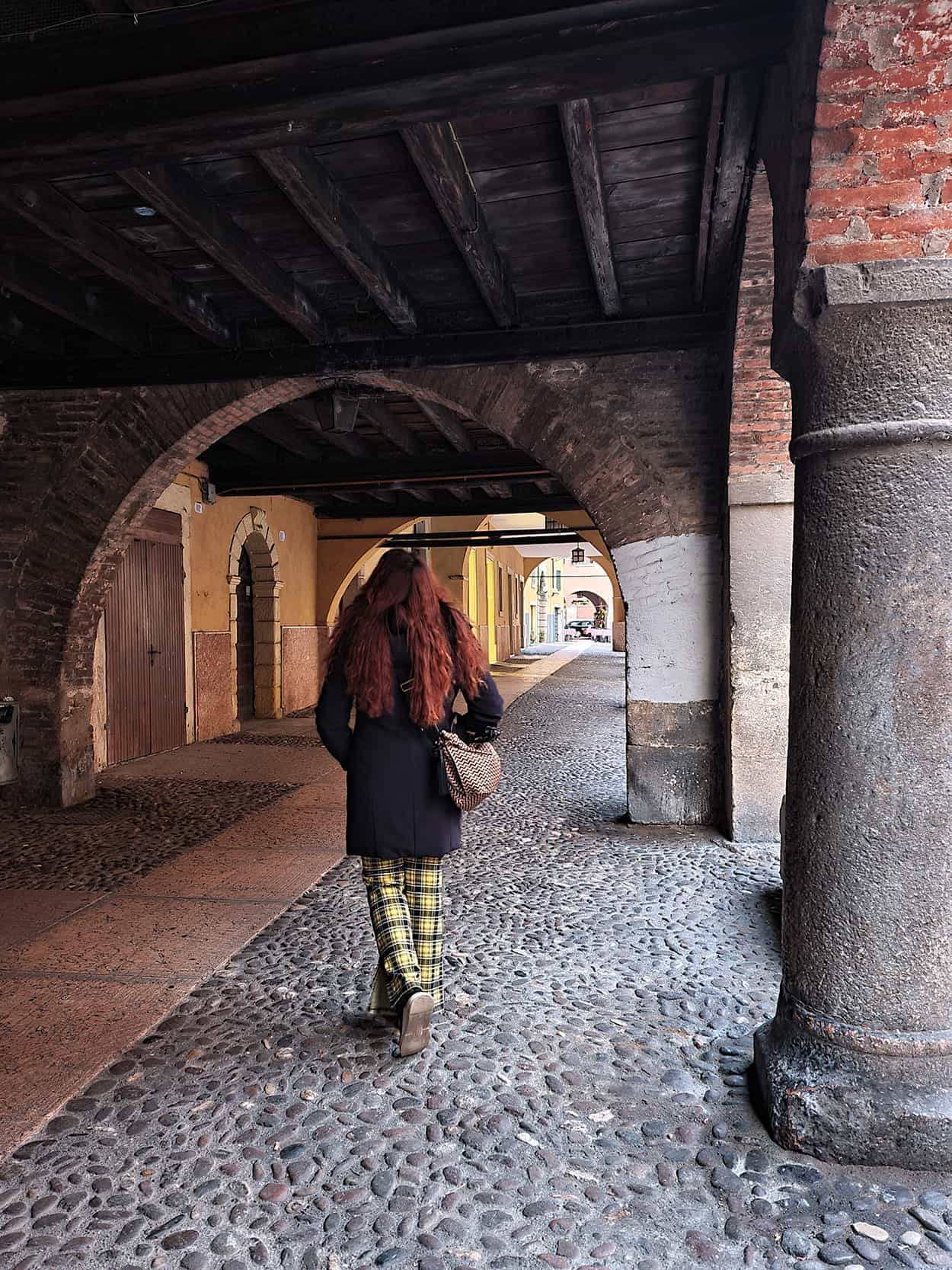 Carlotta di spalle mentre cammina per via sottoriva, un'antica via di Verona che risale al medioevo e che si è perfettamente conservata. E' una strada porticata, con un basso soffitto in legno e con colonne ai lati.