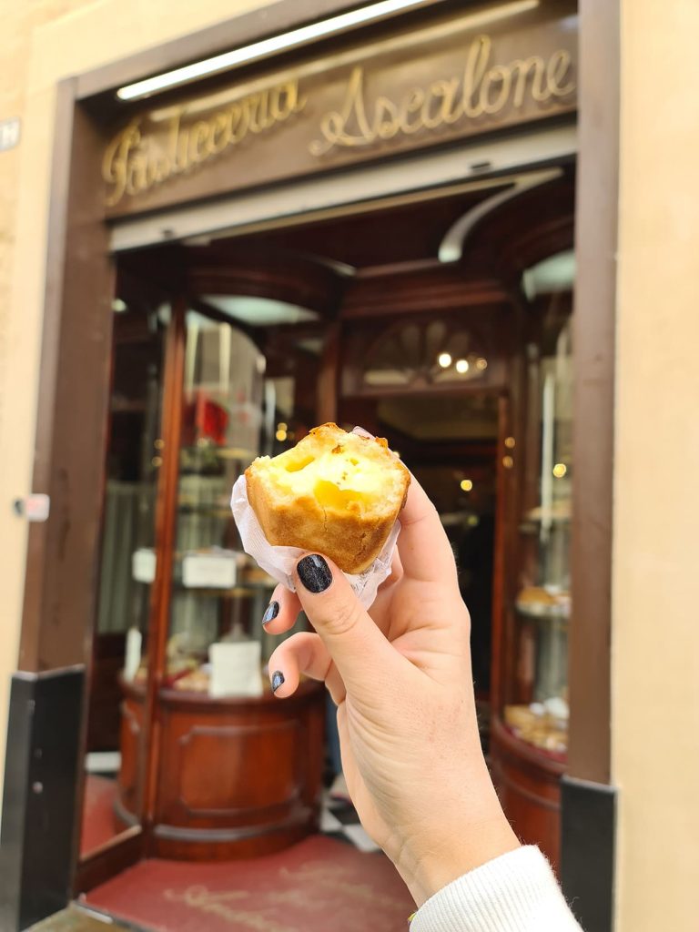 Una foto della mano di Carlotta mentre regge i pasticciotto, dolce tipico del Salento, e sullo sfondo si intravede l'entrata della Pasticceria Ascalone dove è stato inventato.